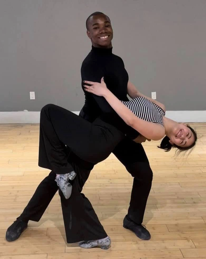 Smiling dance couple performing a dramatic dip in a studio with wooden floors, illustrating the difference between salsa and bachata partner styling